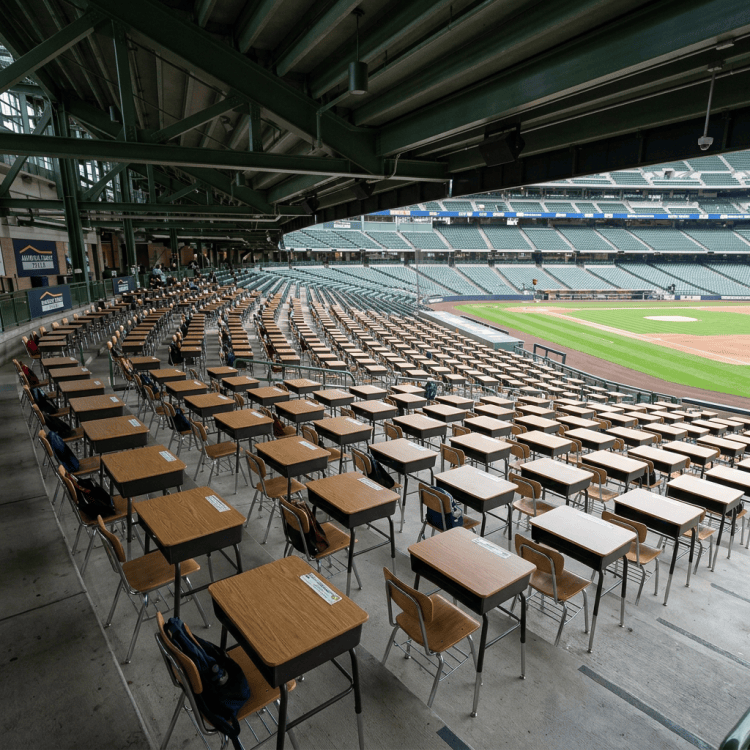 Rows of classroom desks arranged inside a baseball stadium beside the field